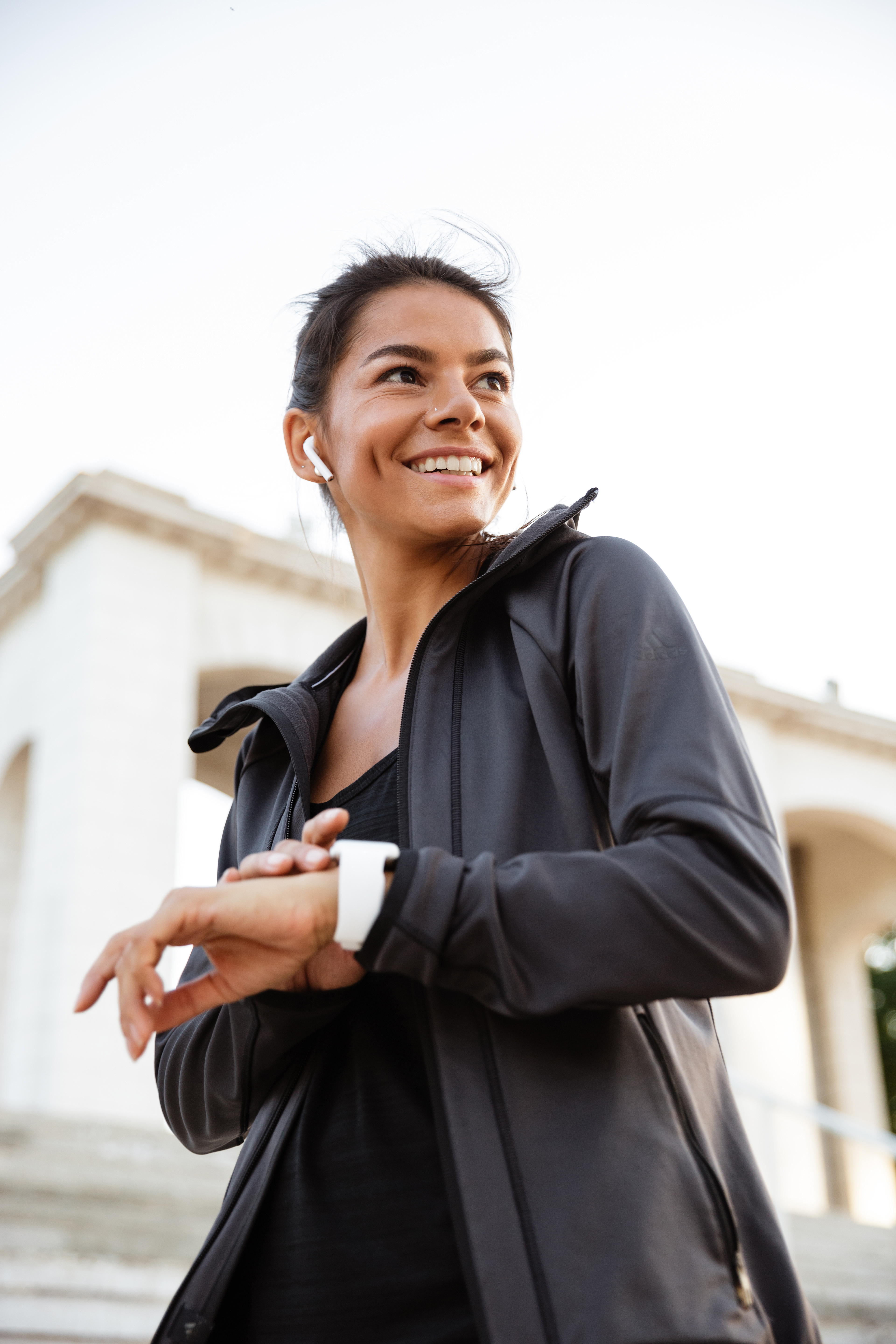 Portrait of a happy fitness woman in earphones checking her smart watch outdoors
